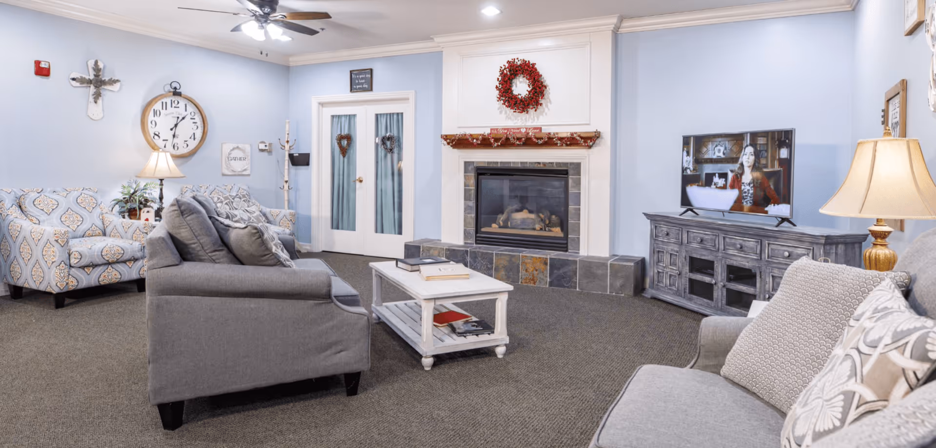A cozy living room in an assisted living facility with light blue walls, a ceiling fan, and a fireplace decorated with a red wreath and garland. The room features a gray sofa, patterned armchairs, a white coffee table with books, a TV on a wooden stand, and lamps providing warm lighting.