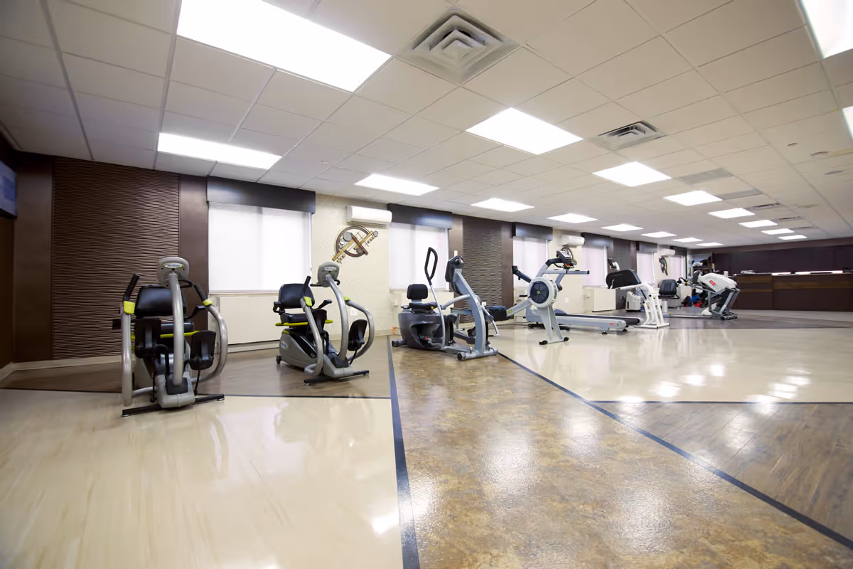 A spacious fitness room in a senior living facility with various exercise equipment including stationary bikes, treadmills, and elliptical machines. The room has a polished floor with different textures and colors, large windows with blinds, and bright overhead lighting.
