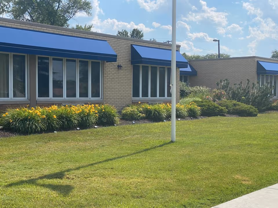 Exterior view of a single-story brick building with blue awnings over the windows, surrounded by well-maintained green grass, yellow flowers, and shrubs under a partly cloudy sky.
