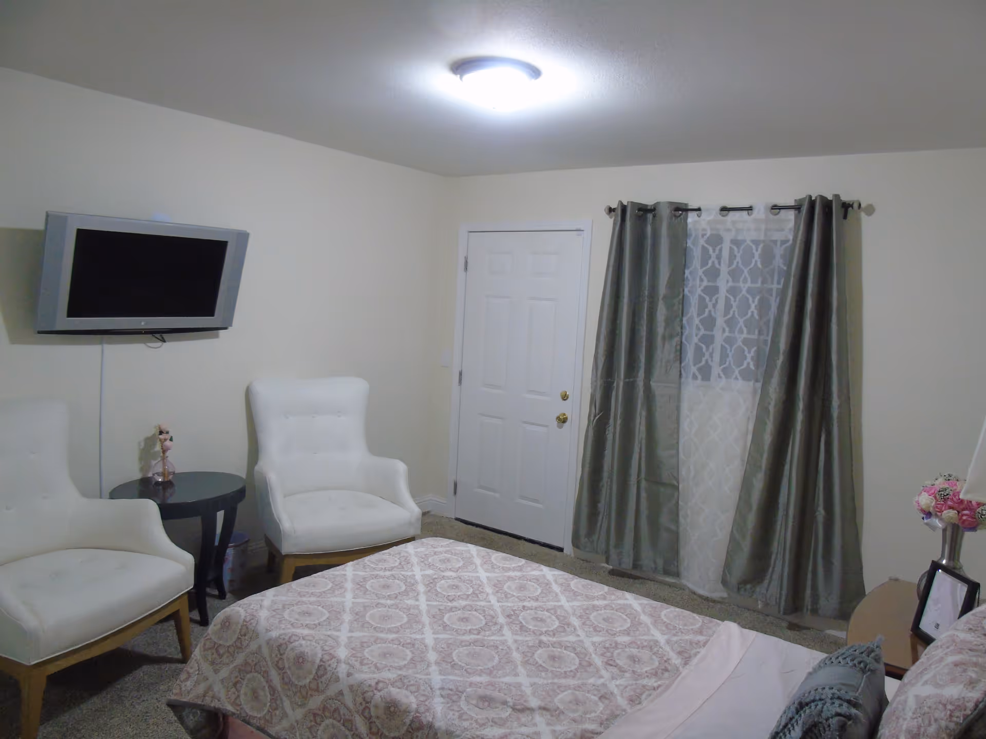 A bedroom with a bed covered in a patterned bedspread, two white armchairs next to a small round table, a wall-mounted flat screen TV, a door, and a window with gray curtains and a sheer white curtain behind them.