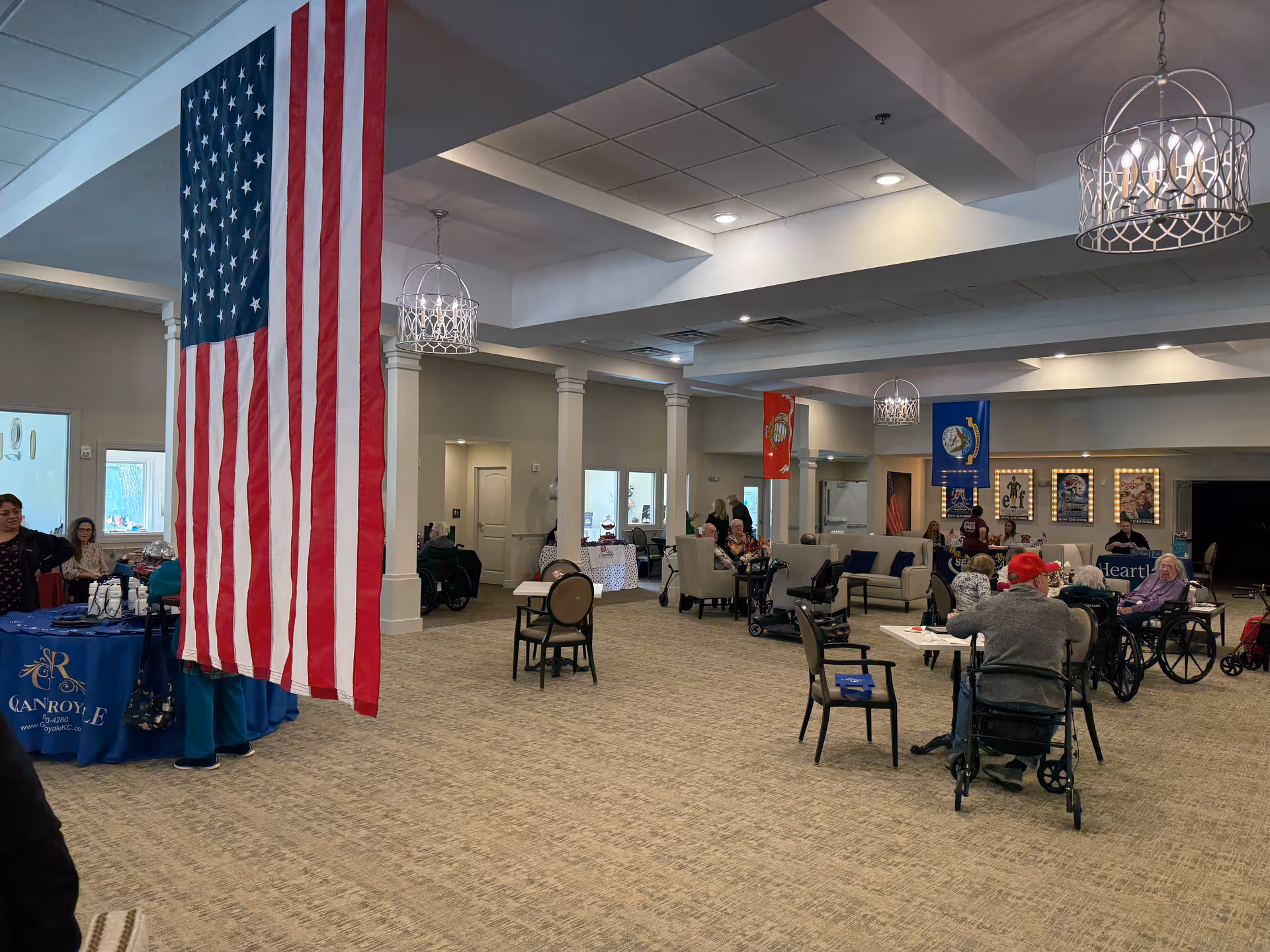 A spacious common area in a senior living facility with several elderly residents seated at tables and in chairs, some in wheelchairs. The room features beige carpeting, white walls, and decorative columns. An American flag hangs prominently from the ceiling. There are banners and posters on the walls, and modern chandeliers provide lighting. A table with a blue tablecloth and some people standing nearby is visible on the left side.