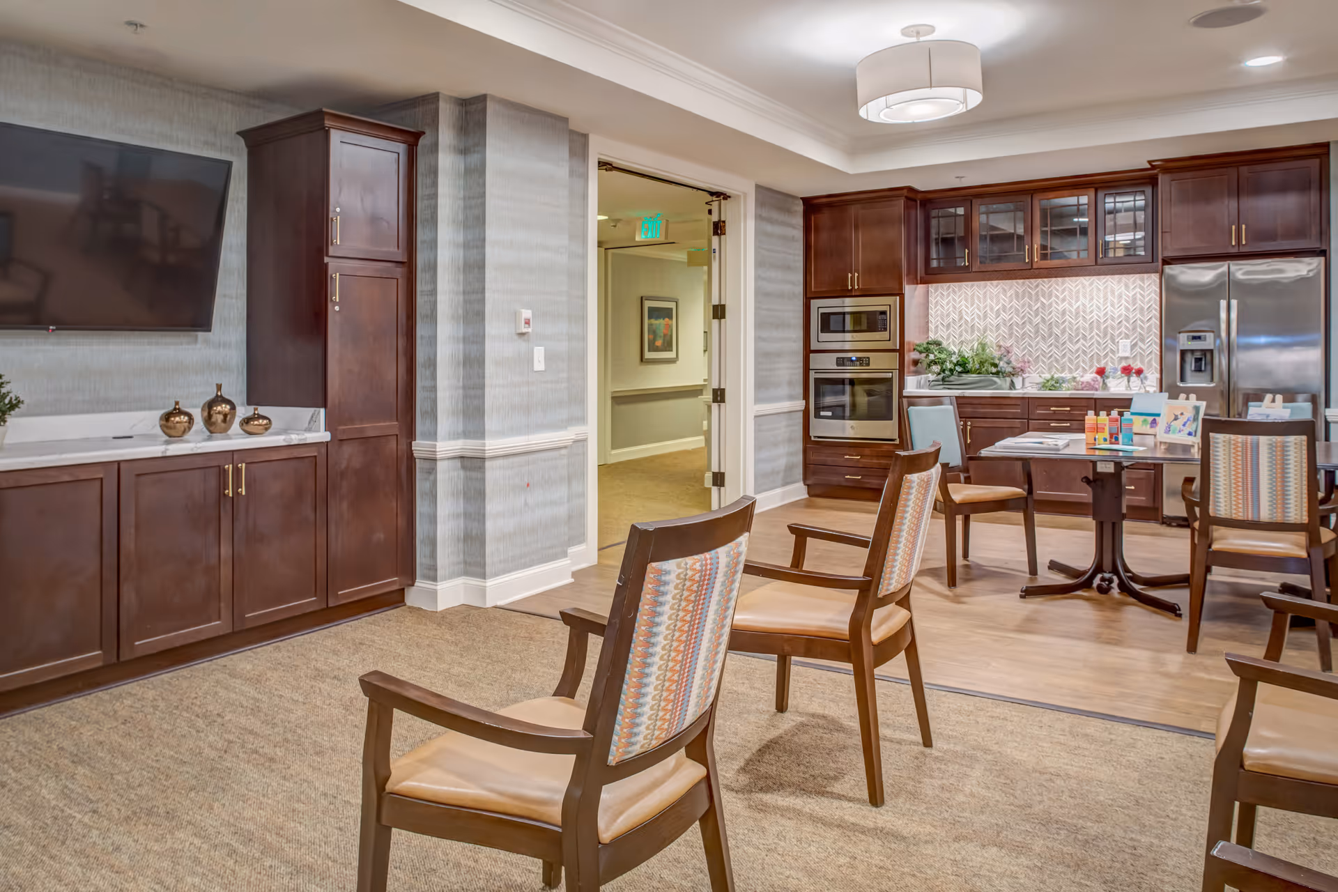 Communal dining and kitchenette area with wooden chairs and tables, a wall-mounted TV, and dark wood cabinetry with stainless appliances.