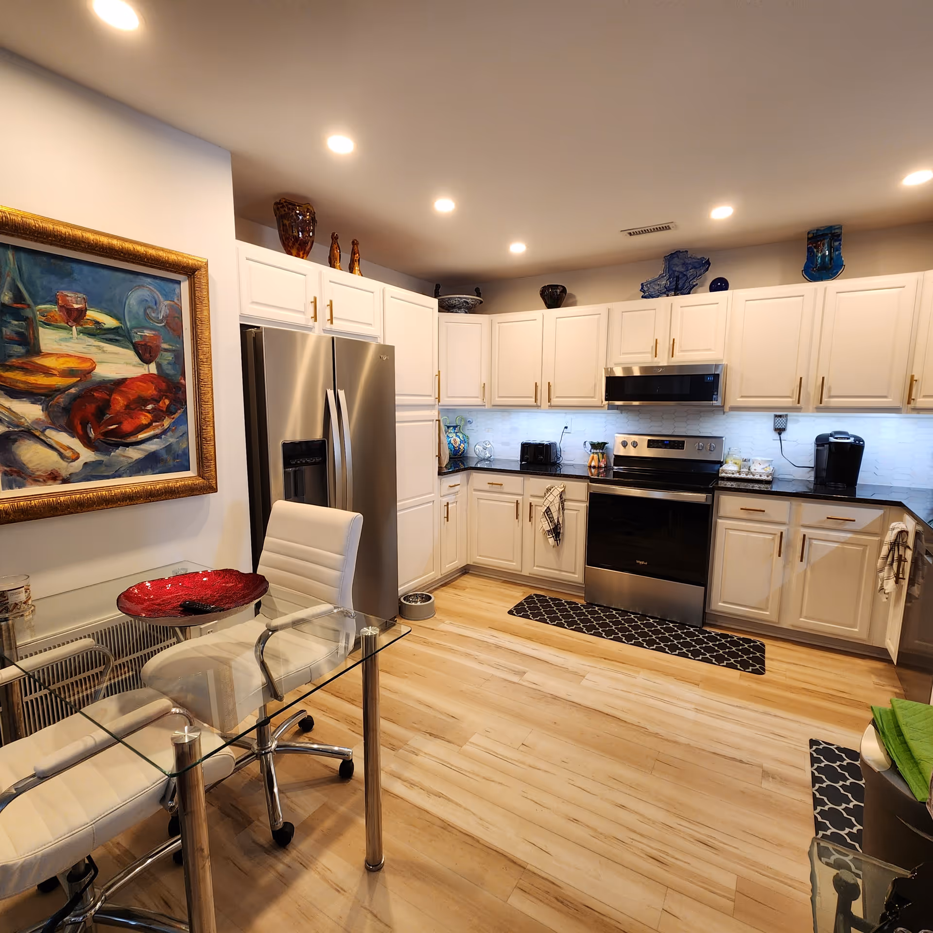 Modern kitchen with white cabinets, stainless steel refrigerator and oven, black countertops, and wooden flooring. There is a glass table with white chairs in the foreground and a colorful painting on the wall to the left. Various decorative items are placed on top of the cabinets.