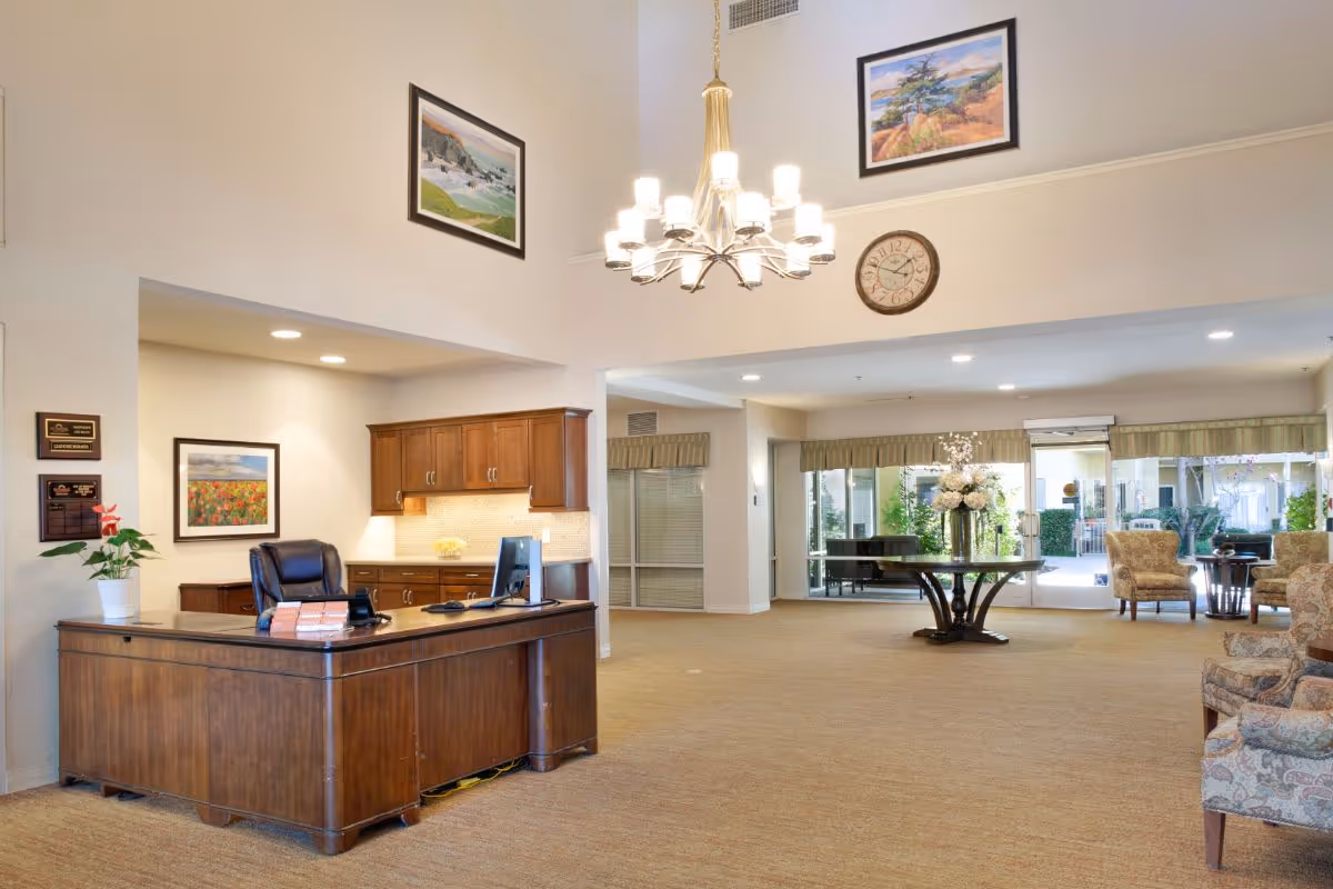 A spacious and well-lit reception area in a senior living facility with a wooden reception desk, a black office chair, and a computer. The room features beige carpeting, a chandelier hanging from a high ceiling, framed landscape paintings on the walls, a round table with a floral arrangement, and several upholstered armchairs. Large windows and glass doors provide a view of an outdoor courtyard with greenery.