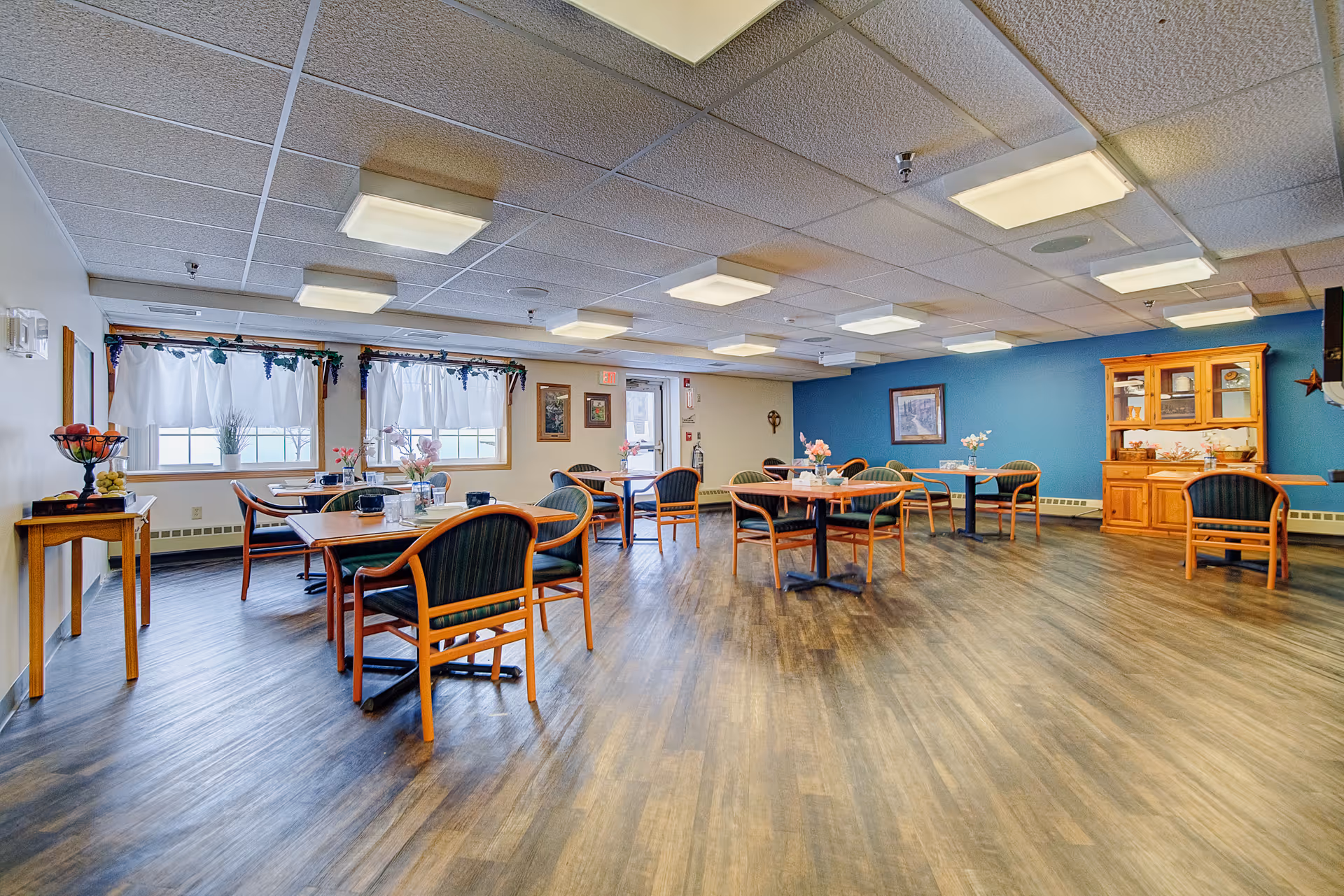 A spacious dining room in an assisted living facility with several wooden tables and chairs arranged neatly. The room has large windows with white curtains, a blue accent wall with framed artwork, and a wooden cabinet displaying dishes. The floor is wood-patterned, and the ceiling has multiple rectangular light fixtures.