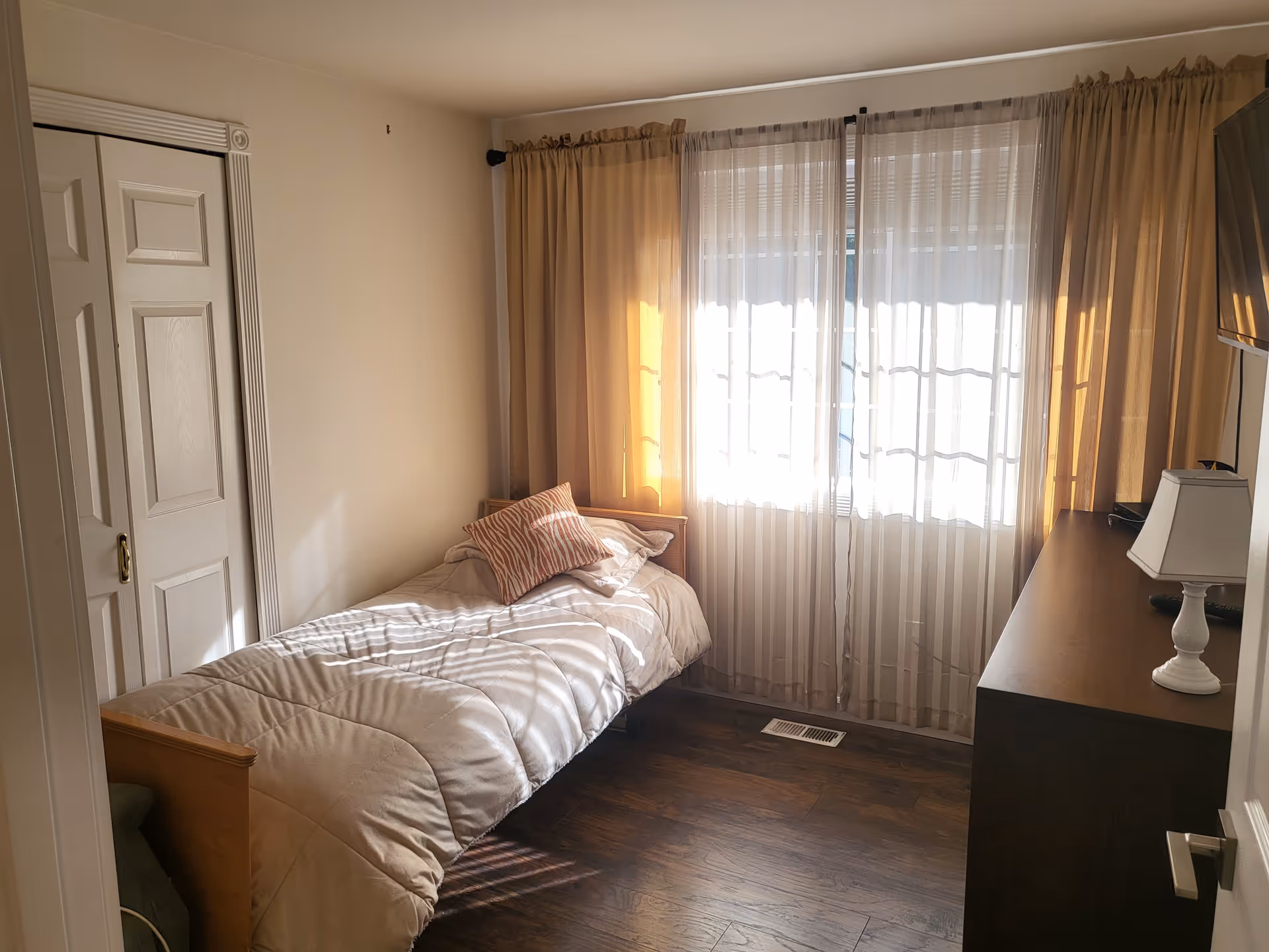 A small bedroom with a single bed covered with a beige comforter and a decorative pillow. There is a window with sheer white curtains and beige drapes letting in sunlight. A wooden dresser with a white lamp and a wall-mounted TV are on the right side. The floor is wooden, and there is a closet with sliding doors on the left.