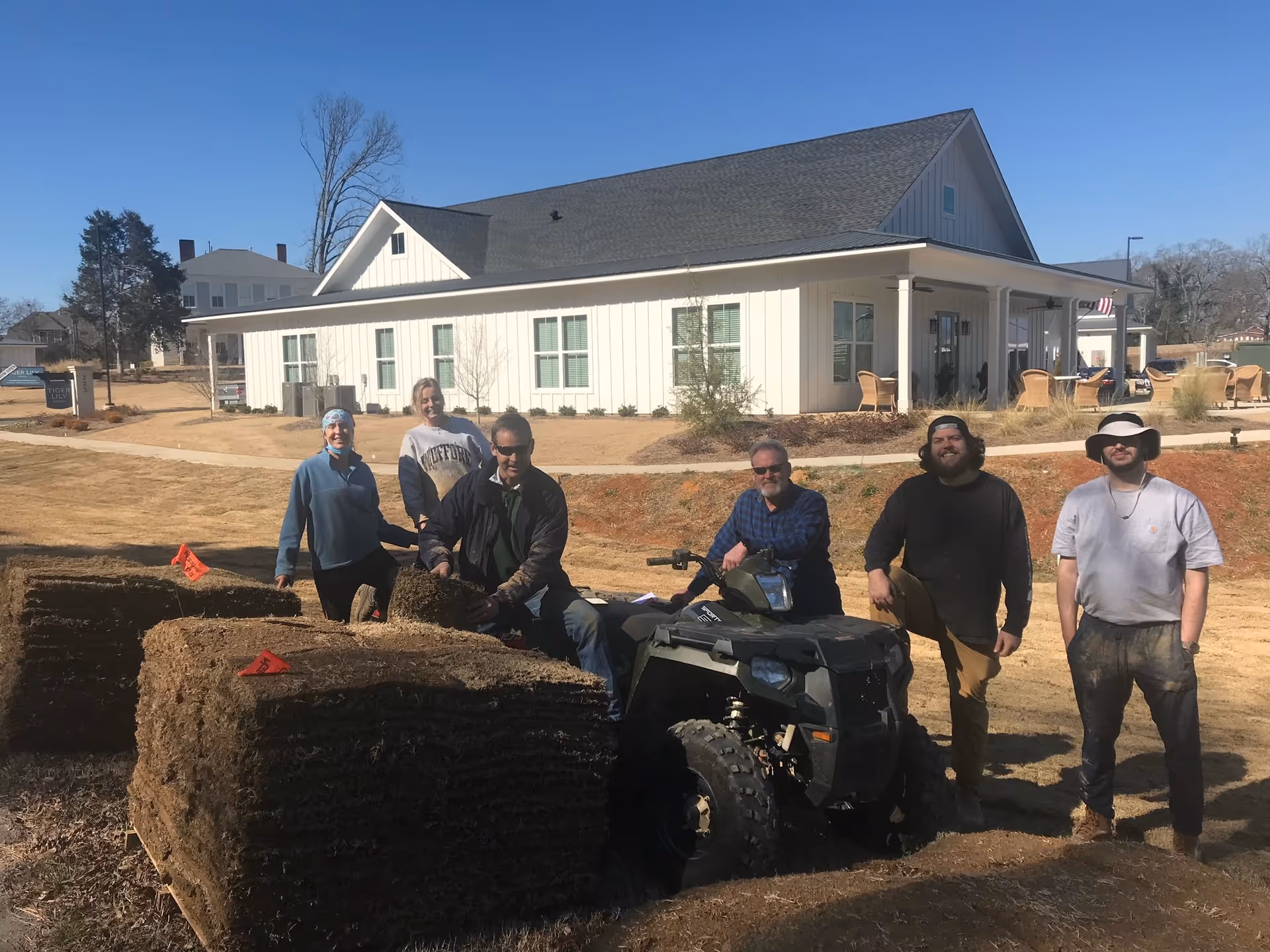 Five people posing outdoors near large stacks of sod rolls and an all-terrain vehicle in front of a white building with a covered porch and outdoor seating under a clear blue sky.