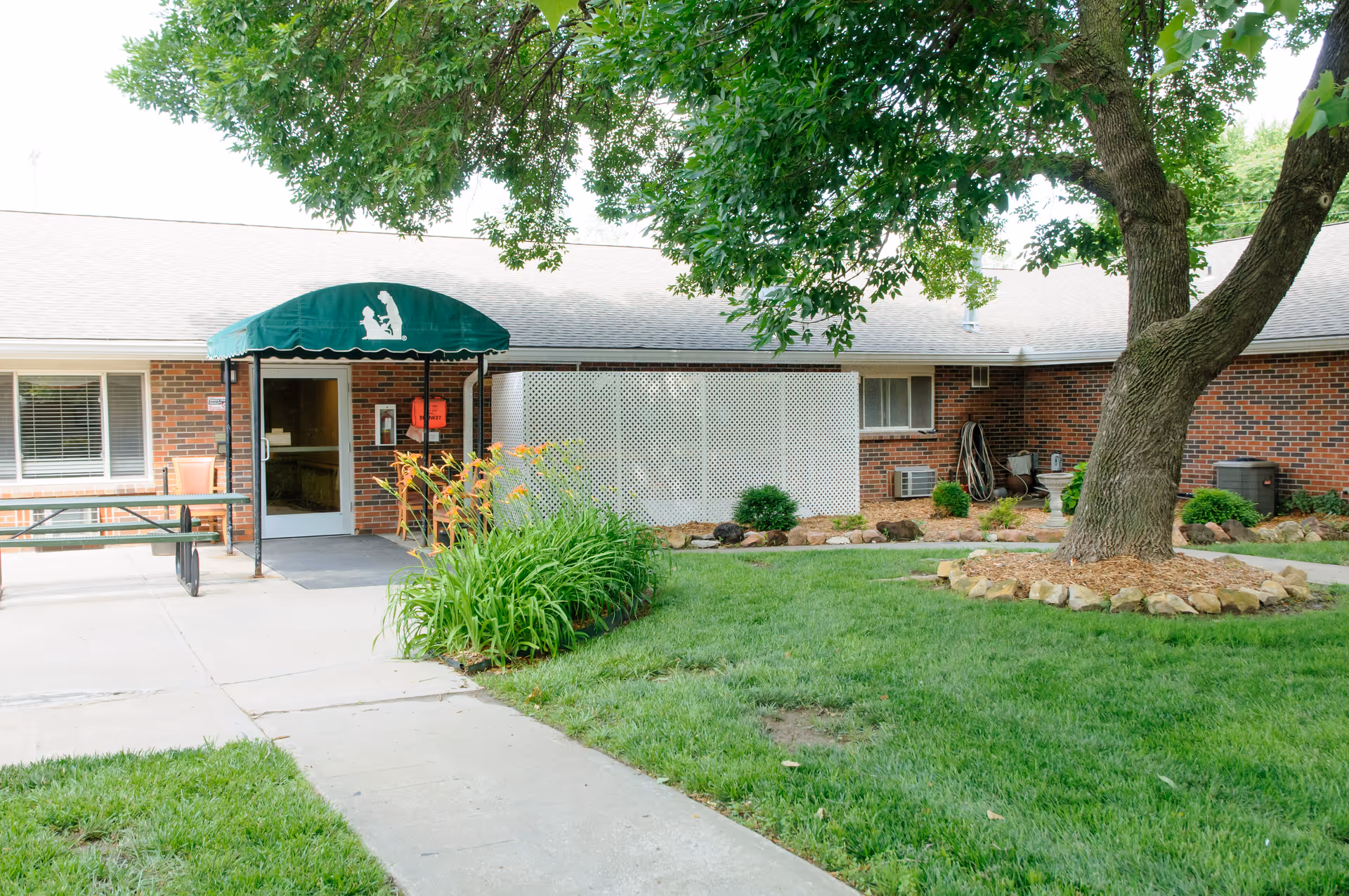 Brick single-story senior living facility entrance with a green awning, sidewalk, landscaping, and a large tree in front.
