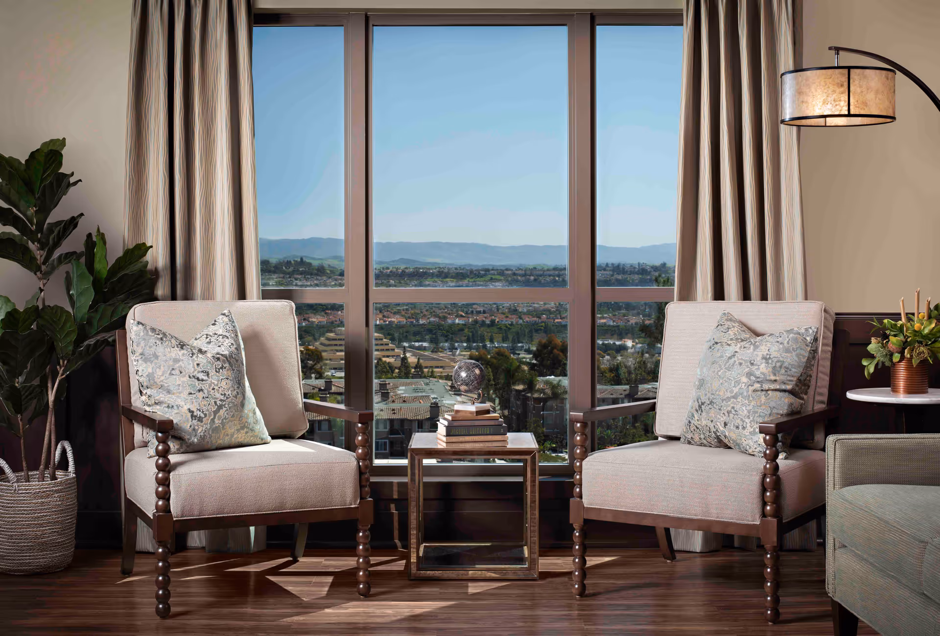 A cozy sitting area with two beige cushioned armchairs featuring wooden legs and armrests, each with a decorative pillow. Between the chairs is a small glass-top side table with a stack of books and a decorative globe. Behind the chairs is a large window with a view of a cityscape and distant mountains under a clear blue sky. Beige curtains frame the window, and a tall green plant in a woven basket is placed to the left. On the right side, a floor lamp with a beige lampshade and a small round table with a floral arrangement are visible.