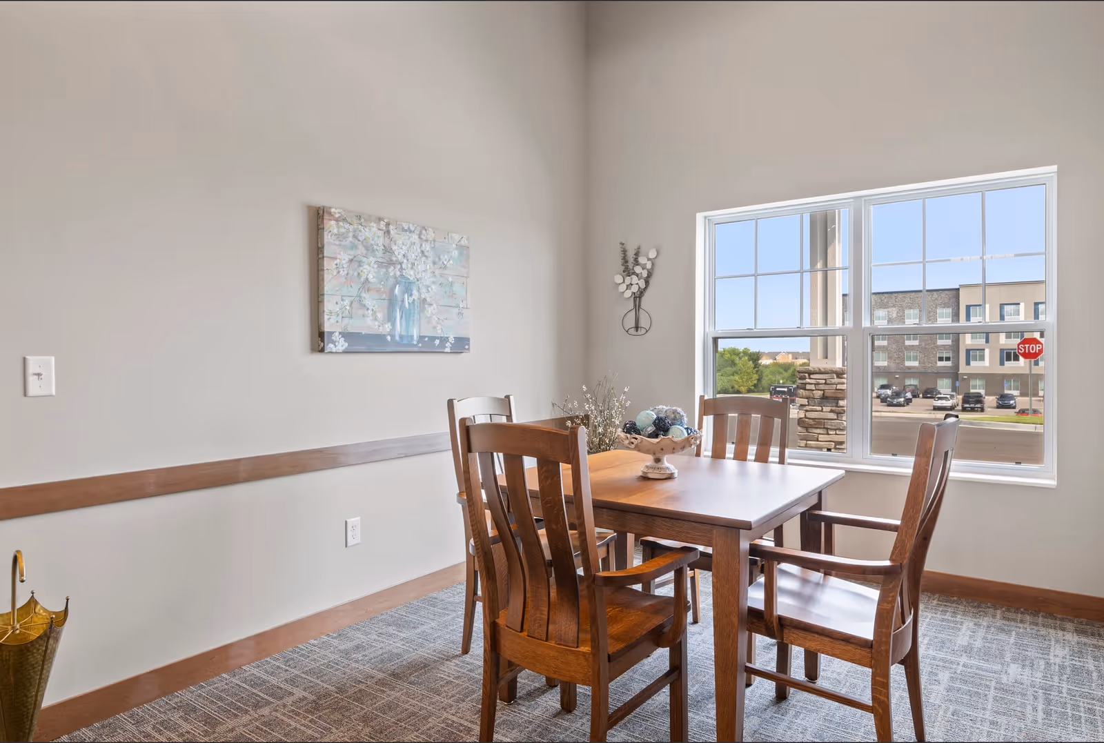 A small dining area with a wooden table and four wooden chairs, two of which have armrests. The table has a decorative bowl with ornamental balls. There is a large window showing a view of a parking lot and a building outside. The walls are light-colored with a floral painting and a wall decoration. The floor is carpeted.