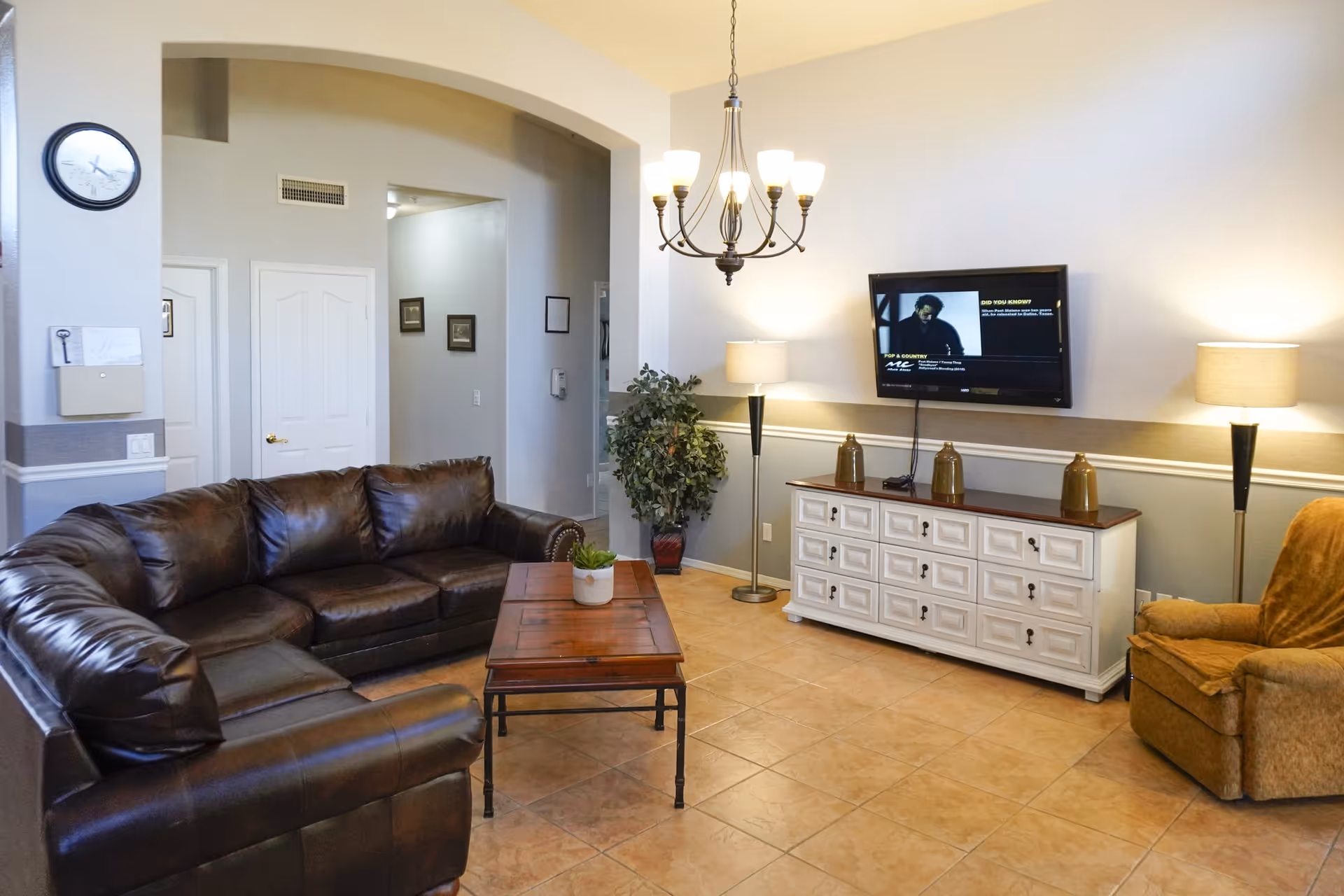 A cozy living room with a dark brown leather sectional sofa, a wooden coffee table with a small plant, a white cabinet with decorative items, a wall-mounted flat-screen TV, two floor lamps, a chandelier, a potted plant, and a single upholstered armchair. The room has tiled flooring and light-colored walls.