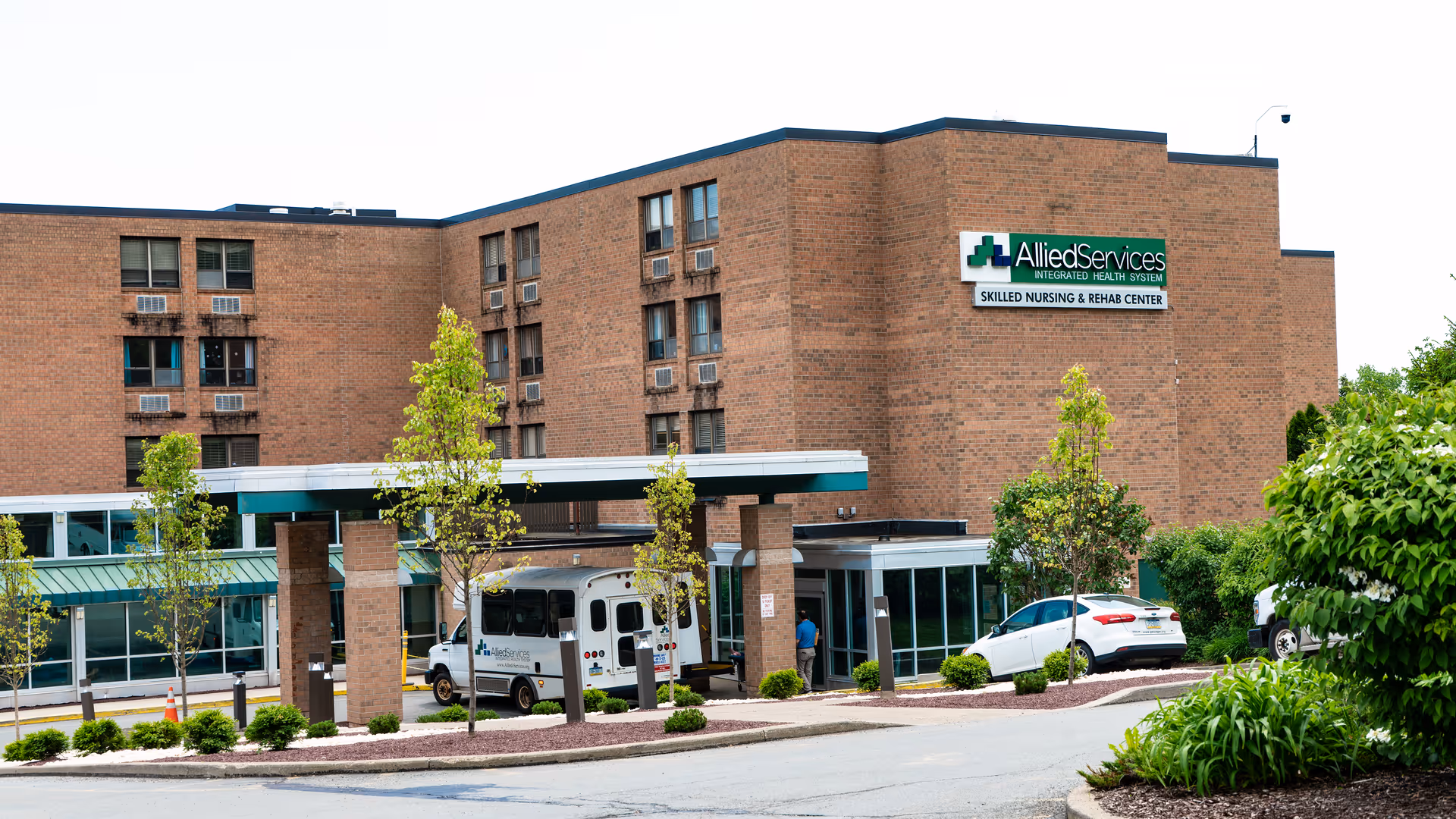 Exterior view of Allied Services Skilled Nursing & Rehabilitation Center, a multi-story brick building with a covered entrance. Several small trees and shrubs are planted around the driveway where a white shuttle bus and a white car are parked.