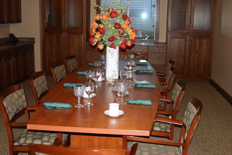 Long wooden dining table set with glassware, green napkins and a tall floral centerpiece in a room with wooden cabinets.