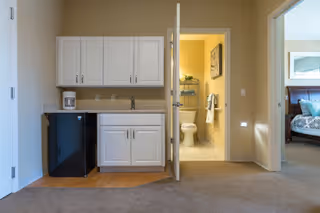 Interior view of a living space in an assisted living facility showing a small kitchenette with white cabinets, a black mini fridge, and a coffee maker. To the right, an open door reveals a bathroom with a toilet and towel rack. Further right, a doorway leads to a bedroom with a bed and pillows.
