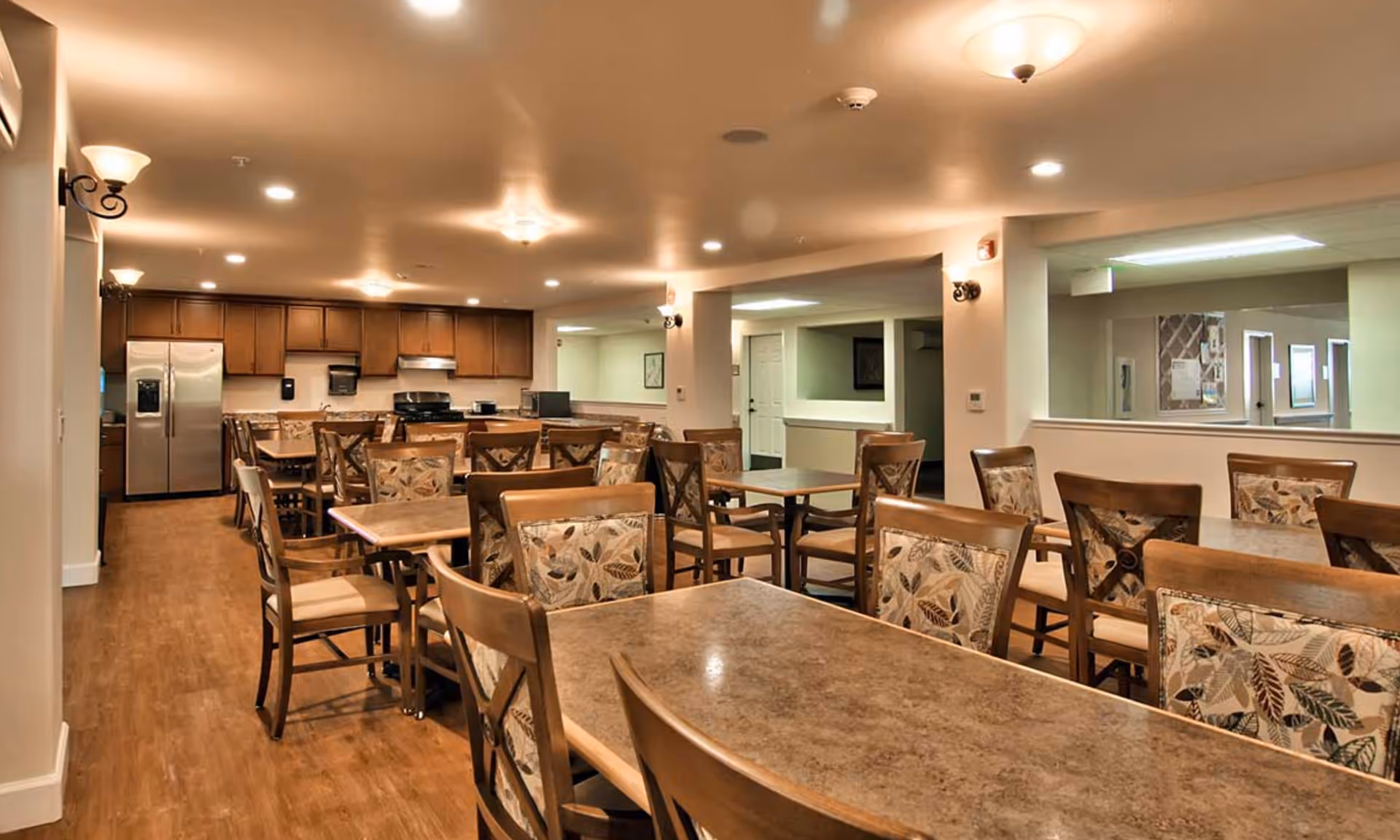 Interior view of a senior living facility dining room with multiple tables and chairs arranged neatly. The chairs have patterned cushions with leaf designs. In the background, there is a kitchen area with wooden cabinets, a stainless steel refrigerator, and a stove. The room has warm lighting and wooden flooring.