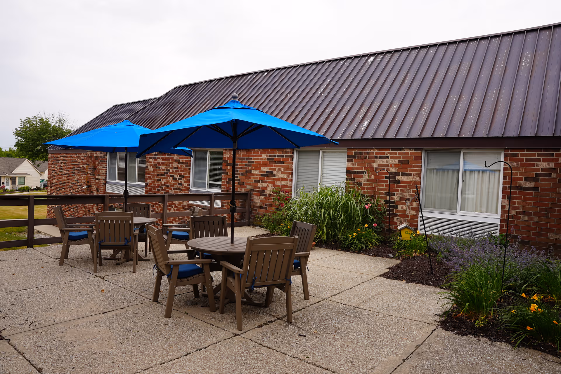 Outdoor patio area with two round tables, each surrounded by four wooden chairs with blue cushions. Each table has a large blue umbrella providing shade. The patio is adjacent to a brick building with windows and some landscaping including green plants and flowers.