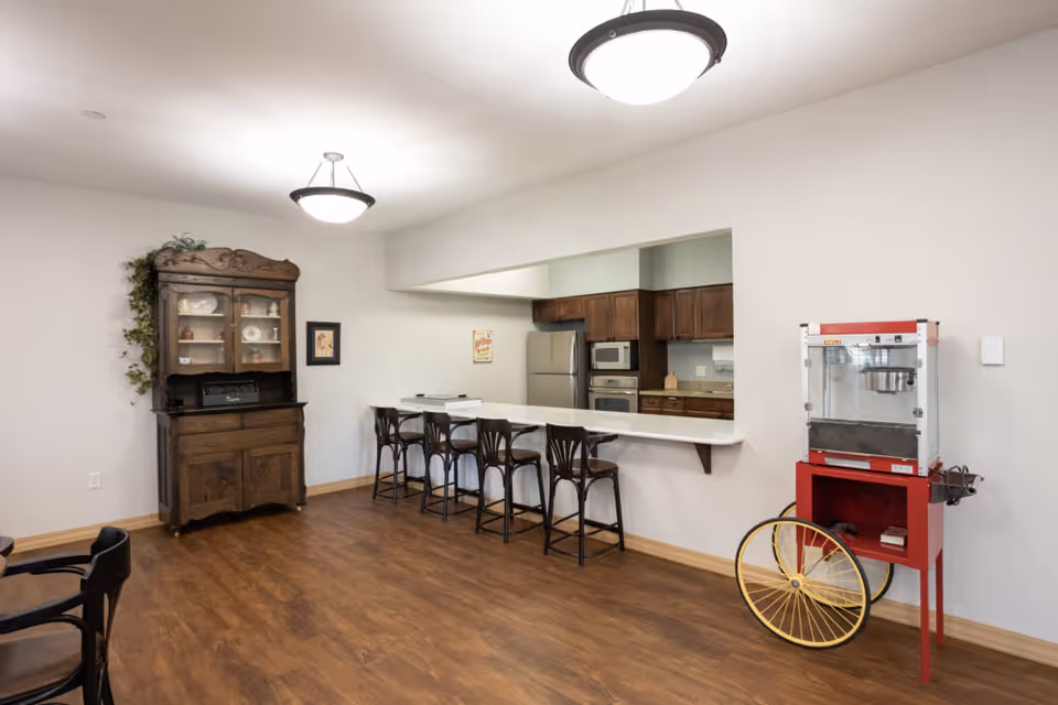 Communal dining area with a long counter and bar stools, kitchenette in the background, an antique hutch, and a popcorn machine on wood floors.