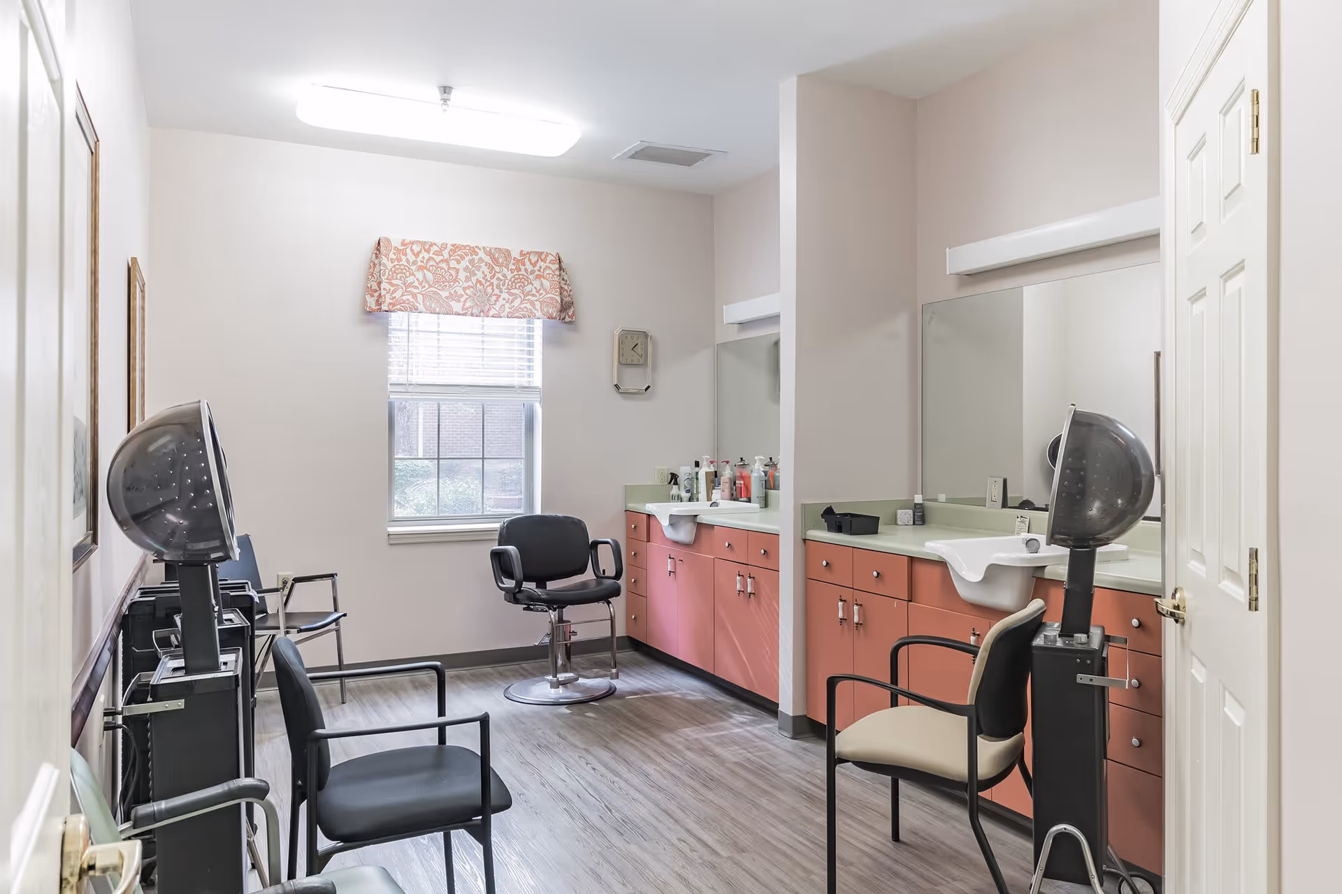 Interior view of a hair salon area in an assisted living facility featuring two hair drying stations with black chairs, a window with a floral valance, pink cabinets with sinks, large mirrors, and additional seating.