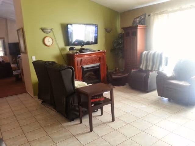 A living room with tiled floor featuring a wall-mounted flat screen TV above a wooden electric fireplace. There are two dark recliner chairs facing the TV, a small wooden side table with books underneath, and two additional chairs near a large window with sheer curtains. A wooden cabinet and a potted plant are in the corner.