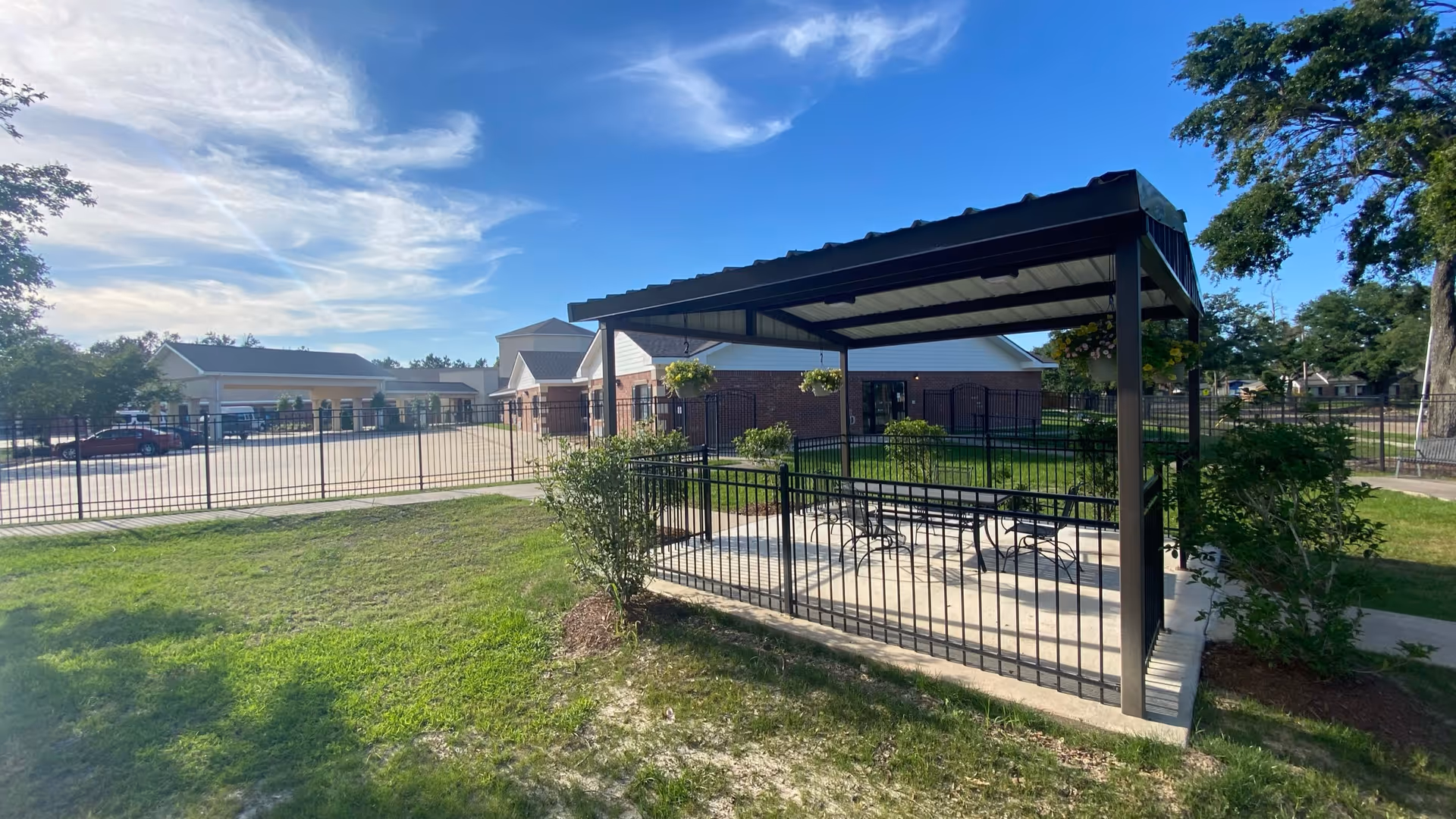 Outdoor view of a senior living facility with a black metal gazebo covering a concrete patio area furnished with tables and chairs. The area is surrounded by a black metal fence, green grass, and some bushes. In the background, there are brick buildings and a parking lot with cars under a partly cloudy blue sky.