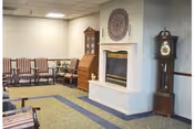 A quiet common area in a senior living facility featuring a fireplace with a decorative wall hanging above it, a grandfather clock to the right, a wooden cabinet with glass doors, and several wooden chairs with striped cushions arranged along the walls.