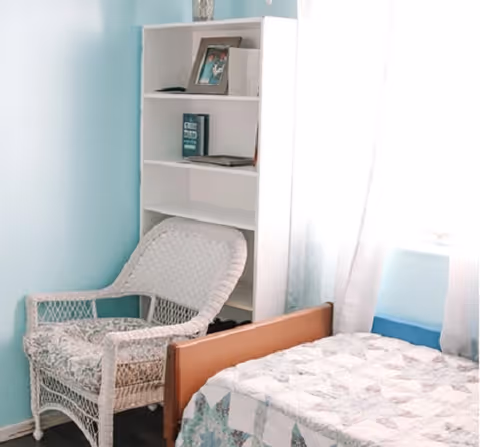Sunlit bedroom corner with a bed, wicker chair, and white bookshelf against a light blue wall.