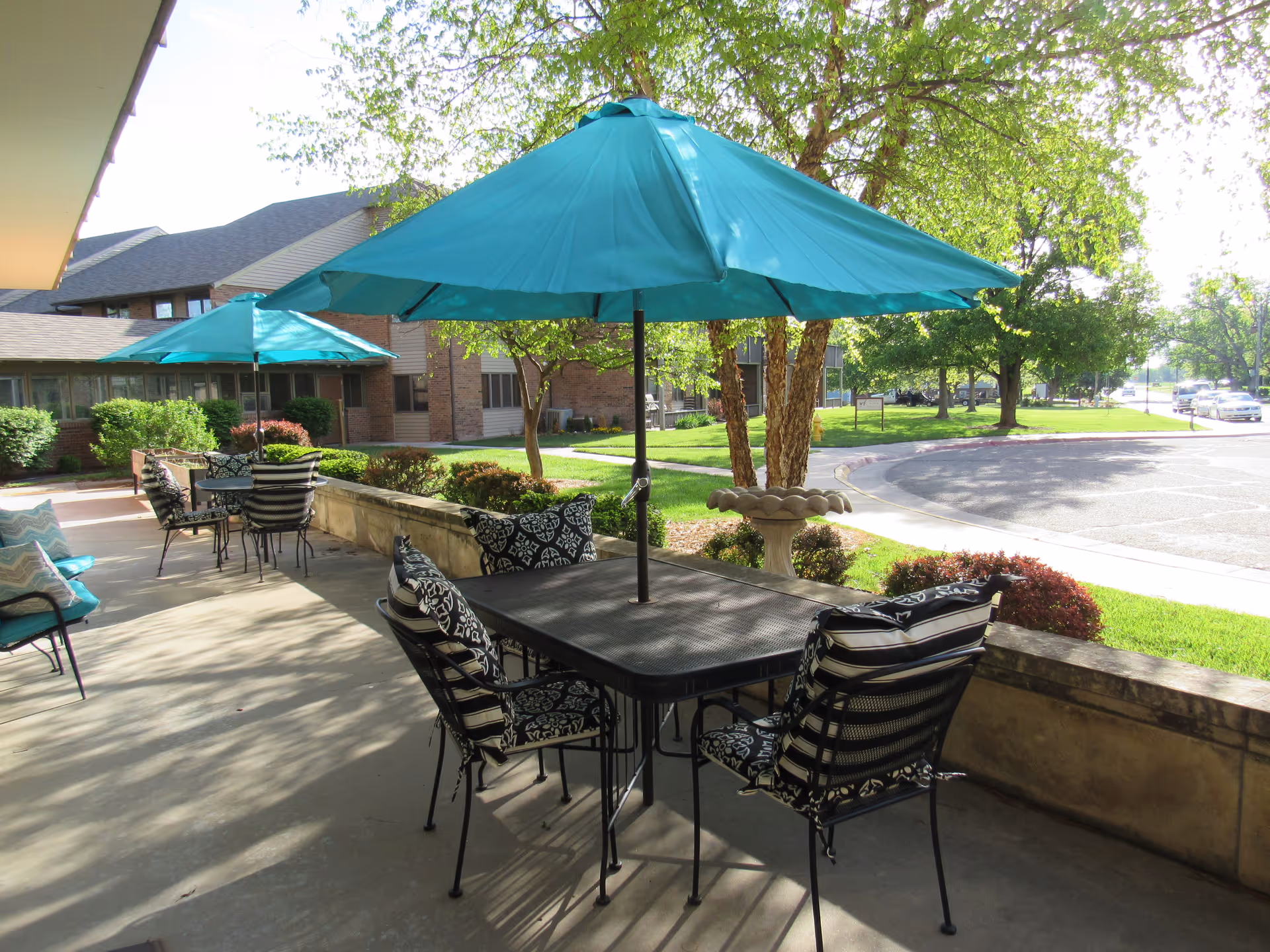 Outdoor patio area with black metal tables and chairs featuring patterned cushions. Each table has a teal umbrella providing shade. The patio overlooks a green lawn with trees and a parking area in the background.