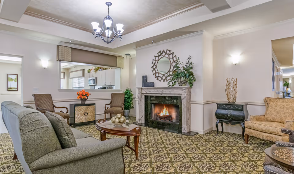 An elegant communal living room with sofas and armchairs arranged around a coffee table and a lit fireplace beneath a decorative mirror.