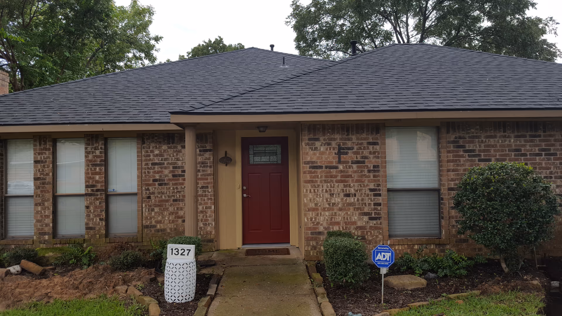 Front exterior view of a single-story brick house with a dark shingled roof, a red front door with a decorative glass panel, two windows on either side of the door with closed blinds, a small garden with bushes and plants, a white decorative mailbox with the number 1327, and a blue ADT security sign in the front yard.