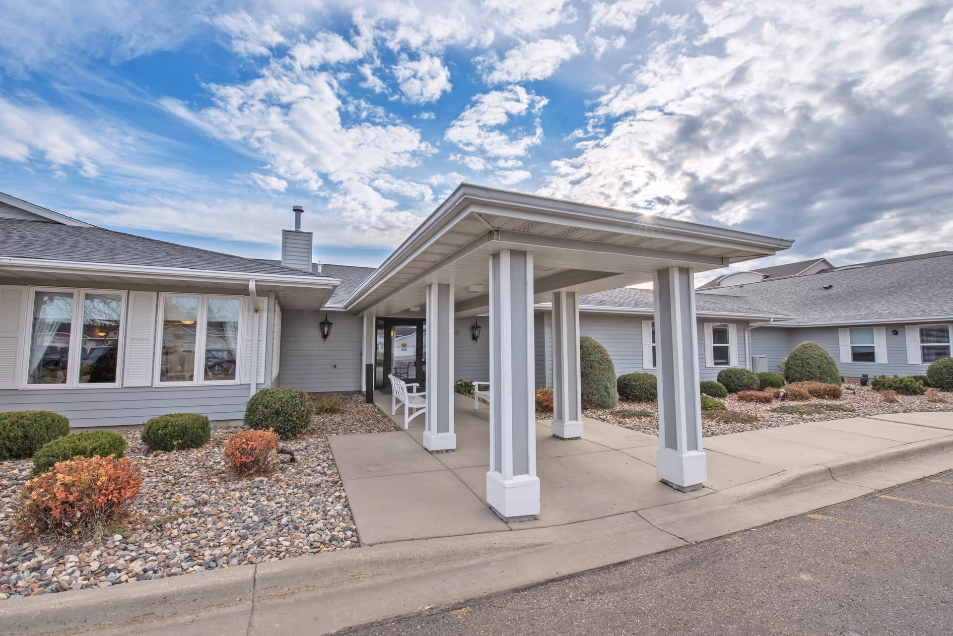 Exterior view of Elison Assisted Living of Minot showing the entrance with a covered walkway supported by white columns, surrounded by landscaped bushes and rocks under a partly cloudy sky.