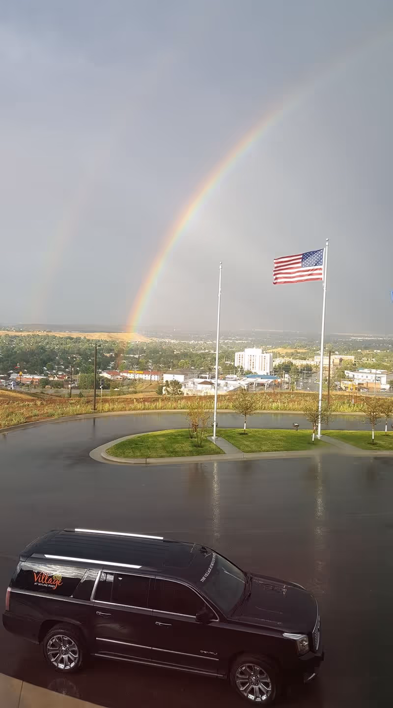 A black SUV with The Village at Skyline Pines logo parked on a wet driveway near a small grassy island with three flagpoles, one of which has an American flag. In the background, there is a cityscape under a cloudy sky with a bright double rainbow.
