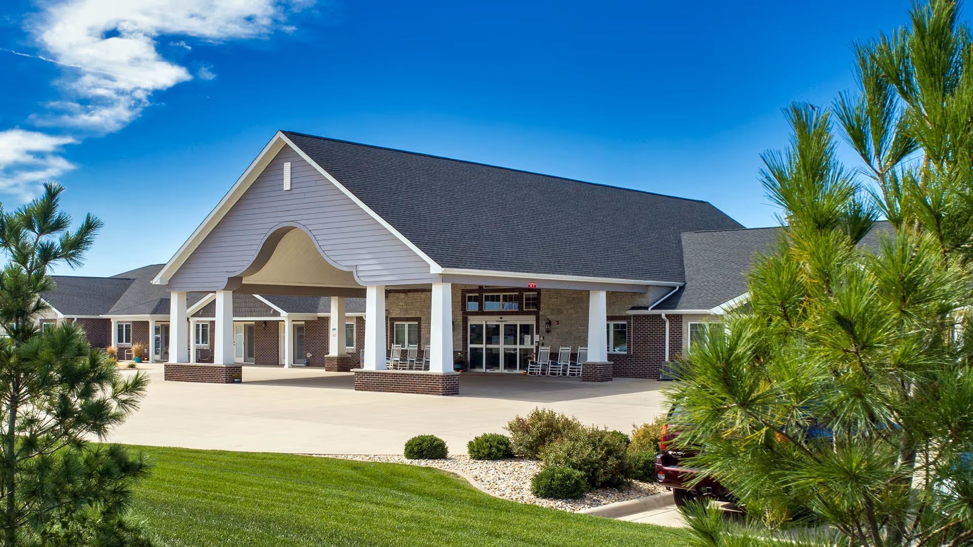 Exterior view of Villas of Holly Brook Assisted Living facility in Washington, IL, showing a large covered entrance with white pillars, brick accents, rocking chairs on the porch, well-maintained green lawn, shrubs, and pine trees under a blue sky with some clouds.
