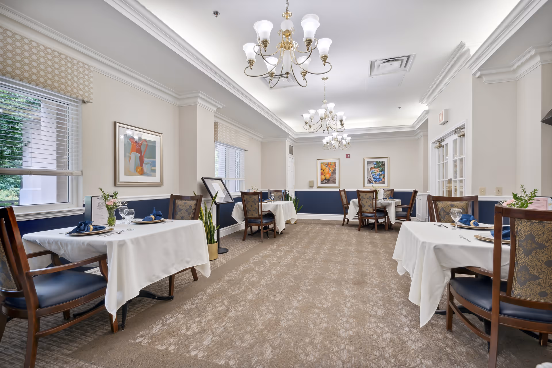 A bright and elegant dining room with several tables covered in white tablecloths, each set with plates, glasses, and napkins. The room features large windows with blinds, framed artwork on the walls, and ornate chandeliers hanging from the ceiling. The carpeted floor and classic wooden chairs with upholstered seats add to the comfortable and refined atmosphere.