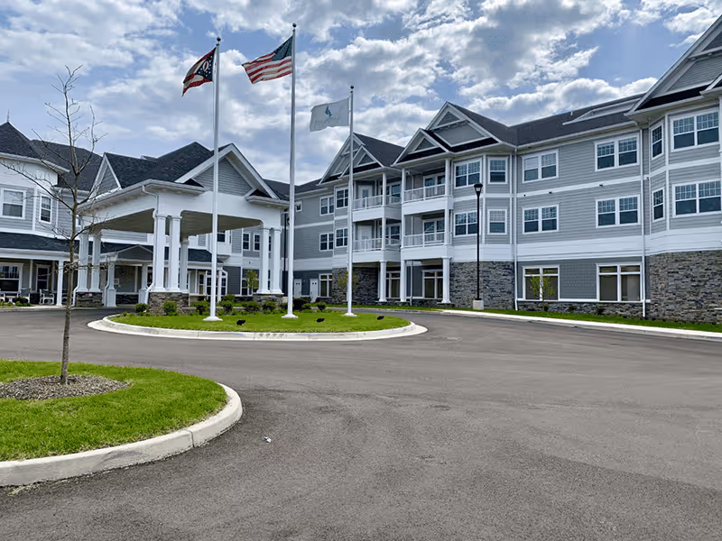 Exterior view of a large senior living facility building with gray and white siding, stone accents on the lower level, and multiple windows. There is a covered entrance with white pillars, a circular driveway with a grassy island in the center, and three flagpoles flying the Ohio state flag, the American flag, and a white flag. The sky is partly cloudy.