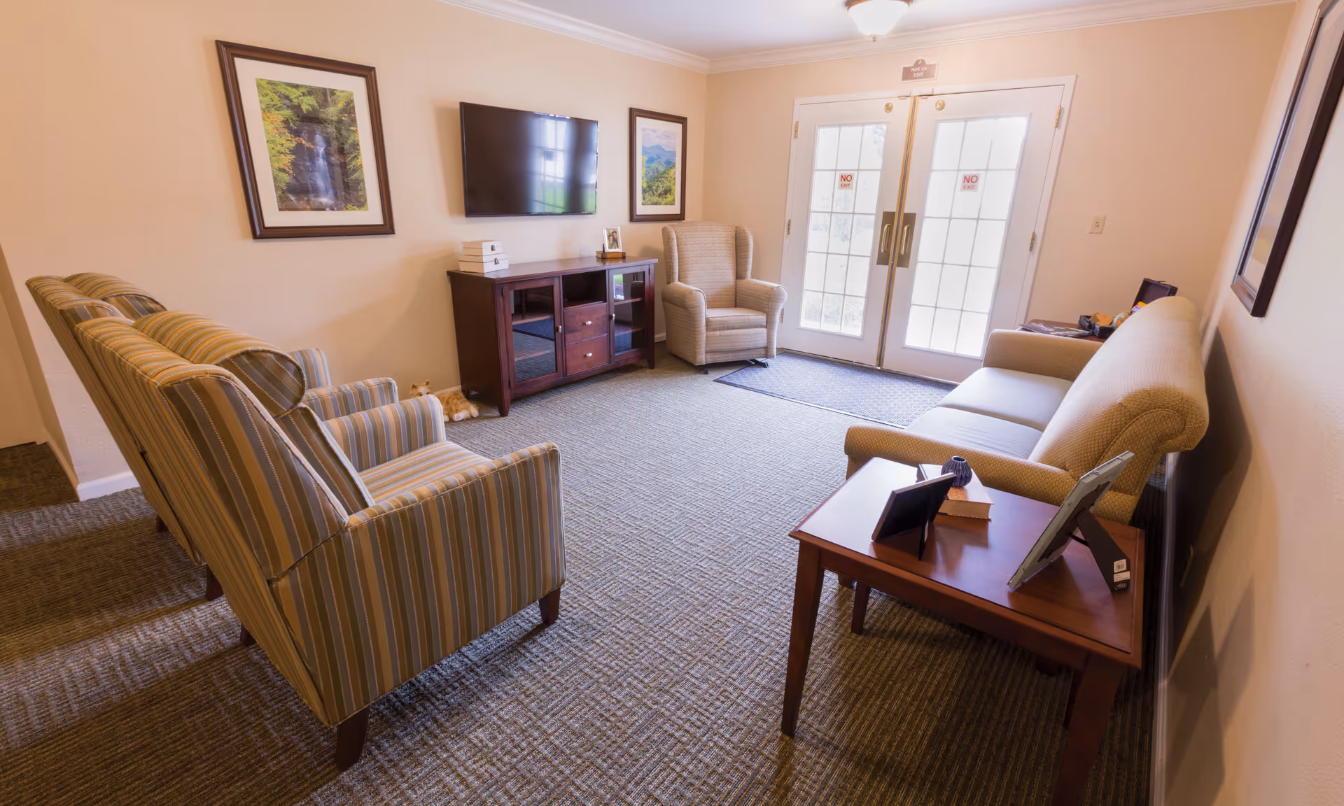 A cozy living room with a beige couch, two striped armchairs, a single beige armchair, a wooden side table with picture frames and a book, a wooden TV stand with a flat-screen TV mounted on the wall above it, and two framed landscape pictures on the wall. There are double glass doors with 'No Exit' signs leading outside.