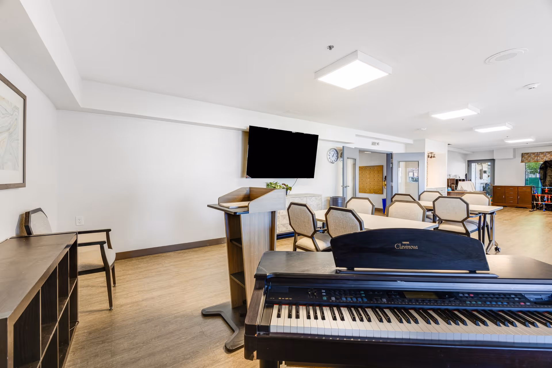 A bright room with a digital piano in the foreground, several chairs and tables arranged in the middle, a podium, a wall-mounted TV, a clock, and a door leading to another room. The floor is wooden, and the walls are white with minimal decoration.