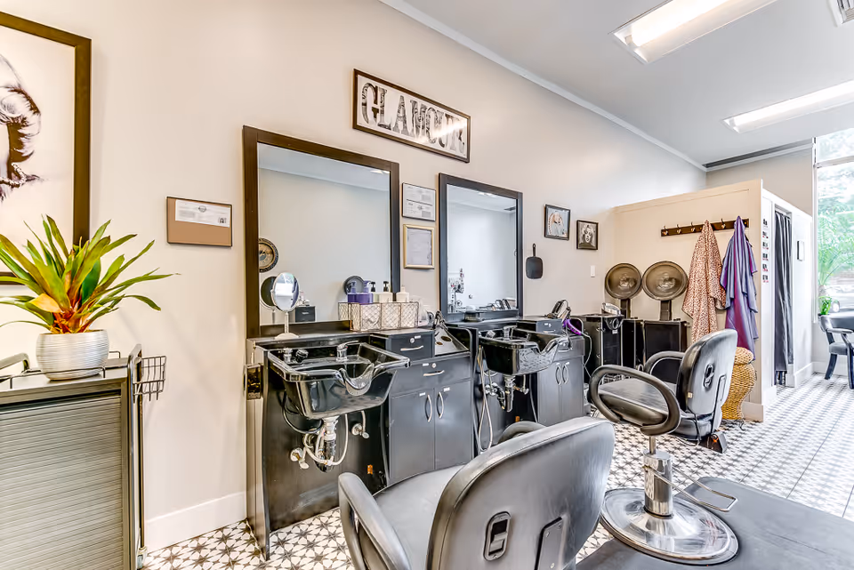 Interior view of a salon area with two black salon chairs in front of black wash basins and mirrors. The room has patterned tile flooring, a plant on a cabinet, and hair drying stations with robes hanging on hooks. The walls are light-colored with framed pictures and a sign that reads 'GLAMOUR'.