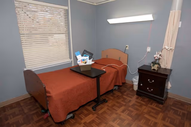 A small, simple bedroom in a senior living facility with a single bed covered in a rust-colored quilt. Next to the bed is a dark wooden nightstand with a small potted orchid and a trash can underneath. A window with closed blinds is on the left wall, and a wall-mounted light fixture is above the bed. A small overbed table holds a basket with various items.
