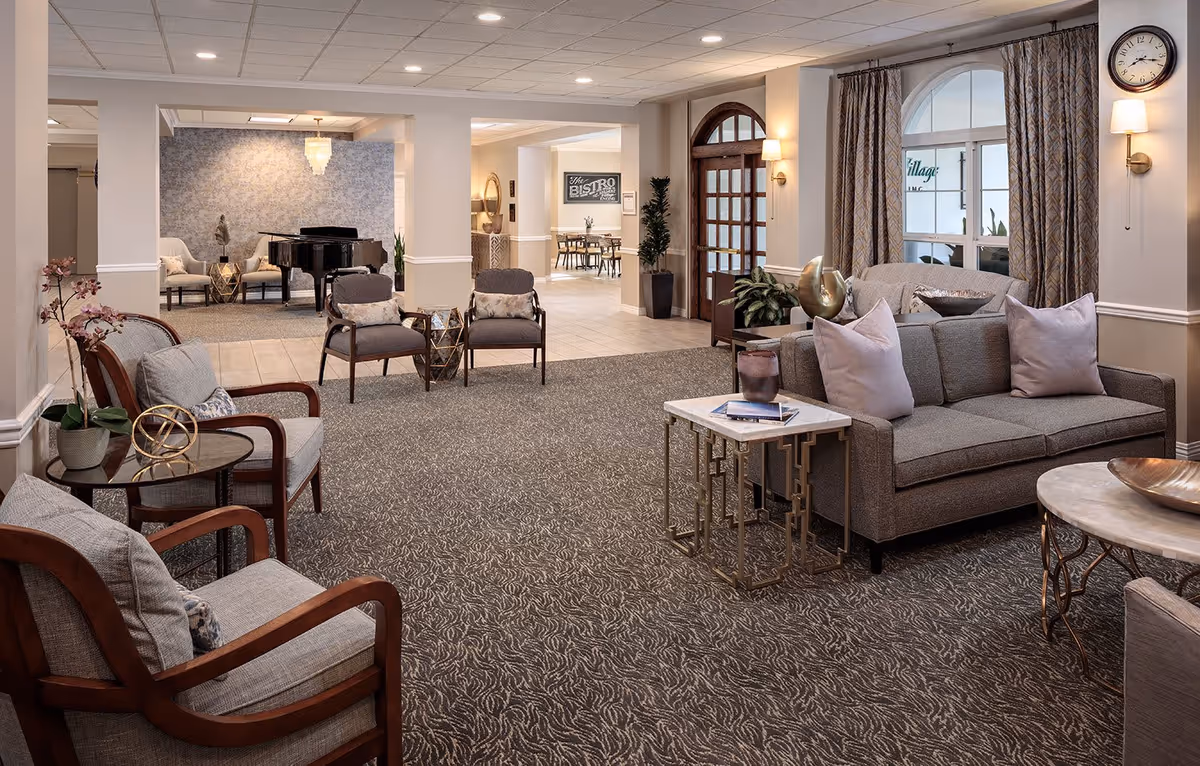 A spacious and elegantly furnished senior living common area with comfortable armchairs and a sofa arranged around small tables. In the background, there is a grand piano and additional seating. The room features soft lighting, patterned carpet, and large windows with curtains. A clock is mounted on the wall near the entrance door.
