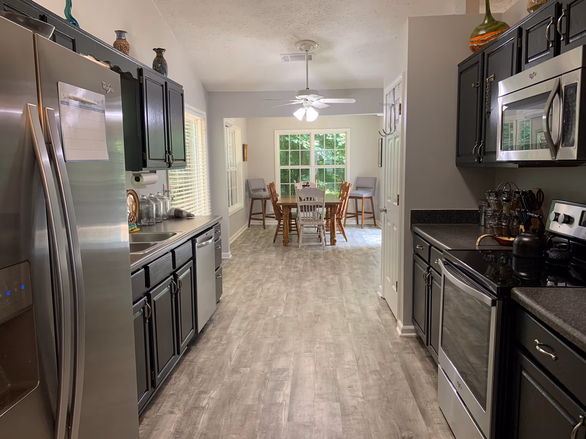 A modern kitchen with stainless steel appliances including a refrigerator, dishwasher, microwave, and stove. The kitchen has dark cabinets and a light wood floor. At the end of the kitchen is a dining area with a wooden table, several chairs, and large windows letting in natural light.