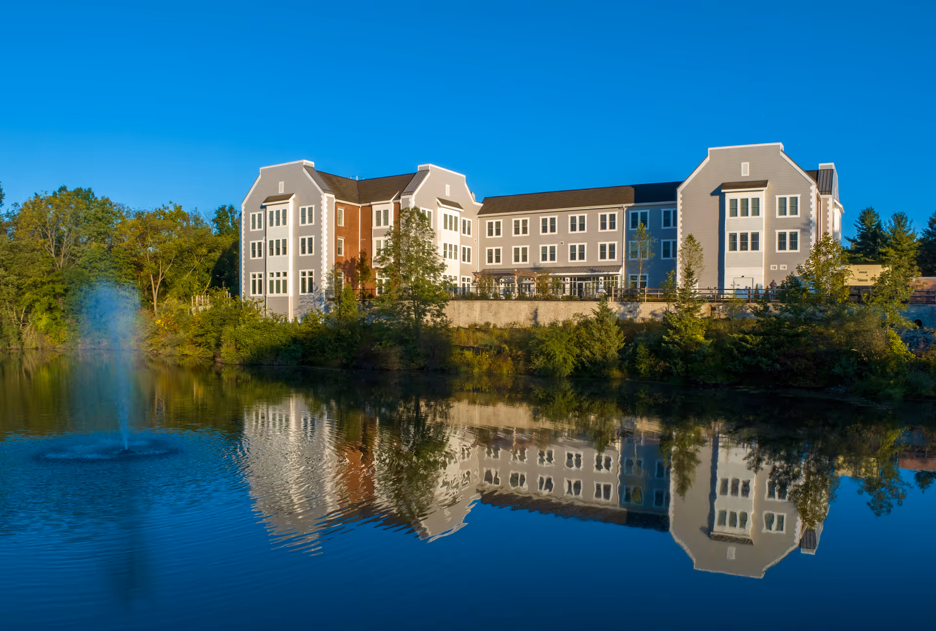 Multi-story residential building reflected in a pond with a fountain and surrounding trees under a clear blue sky.