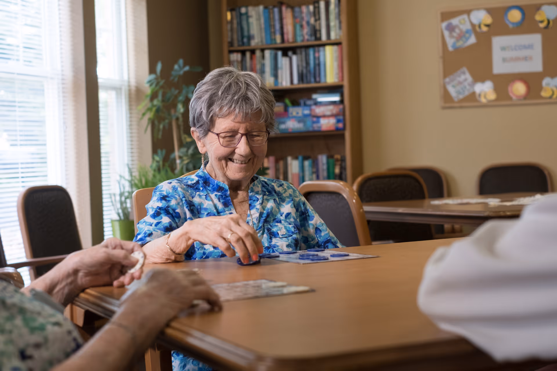 An elderly woman with short gray hair and glasses is smiling while playing a board game at a table in a well-lit room. There are other people partially visible around the table. Behind her, there is a bookshelf filled with books and board games, and a bulletin board with a 'Welcome Summer' sign and decorations.