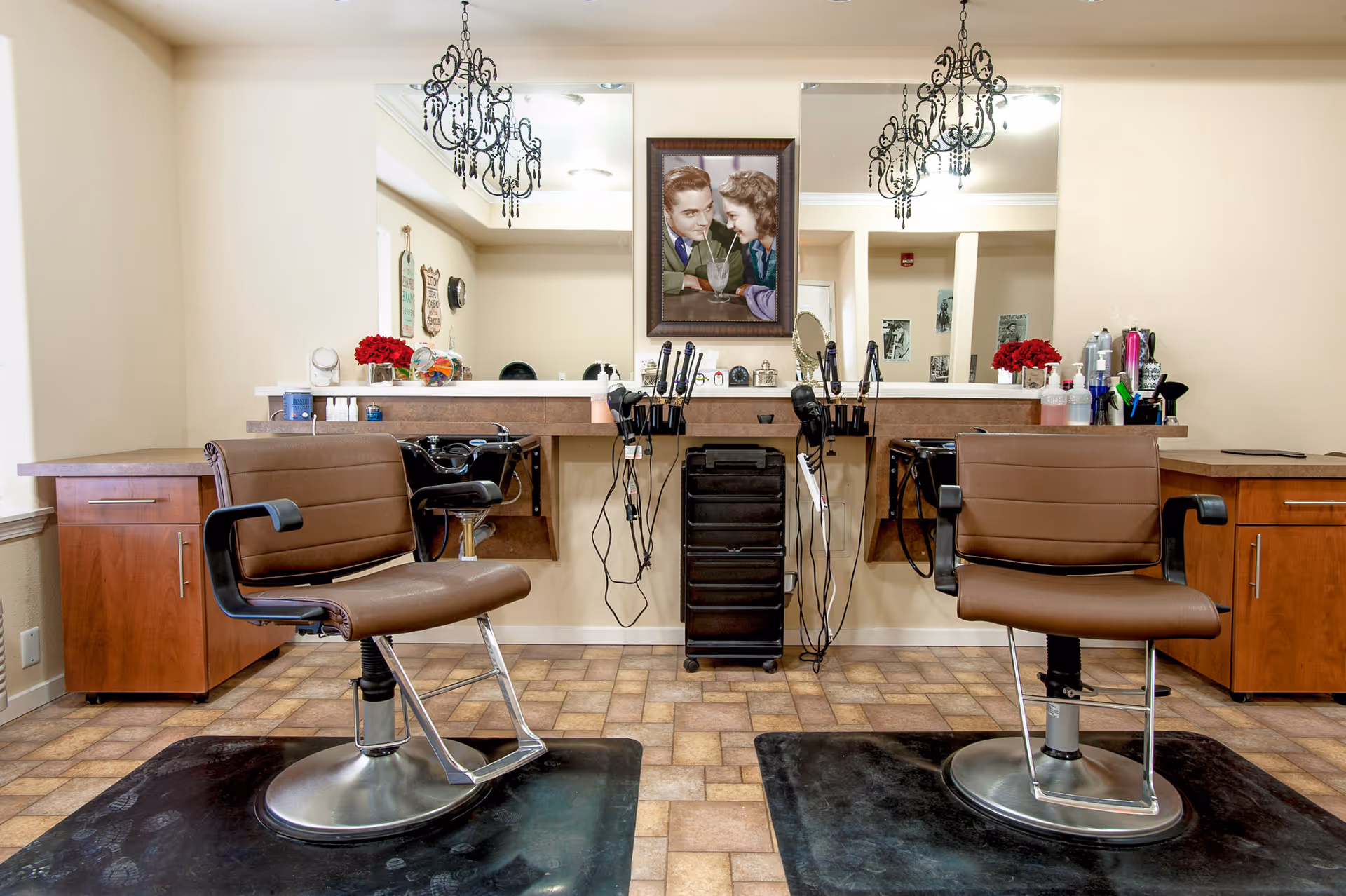 Interior view of a hair salon area with two brown salon chairs on black mats facing a counter with two sinks and mirrors. The counter has various hair styling tools and products. Above the counter is a framed vintage-style picture of a man and woman sharing a milkshake. Two black chandeliers hang from the ceiling.