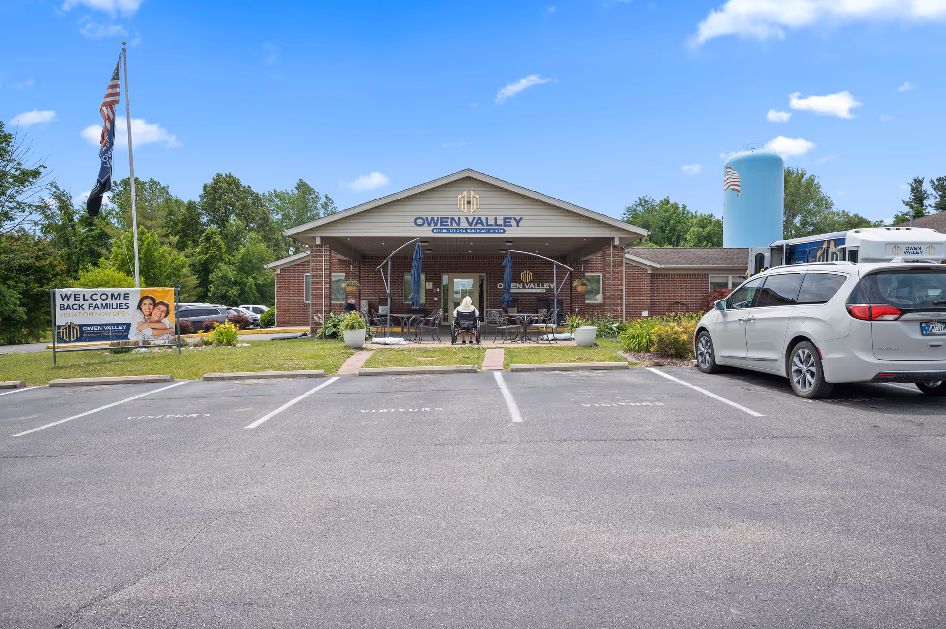 Front entrance of Owen Valley Rehabilitation and Healthcare Center showing the brick building, parking spaces, a welcome sign, flagpole, and a person seated on the covered porch.