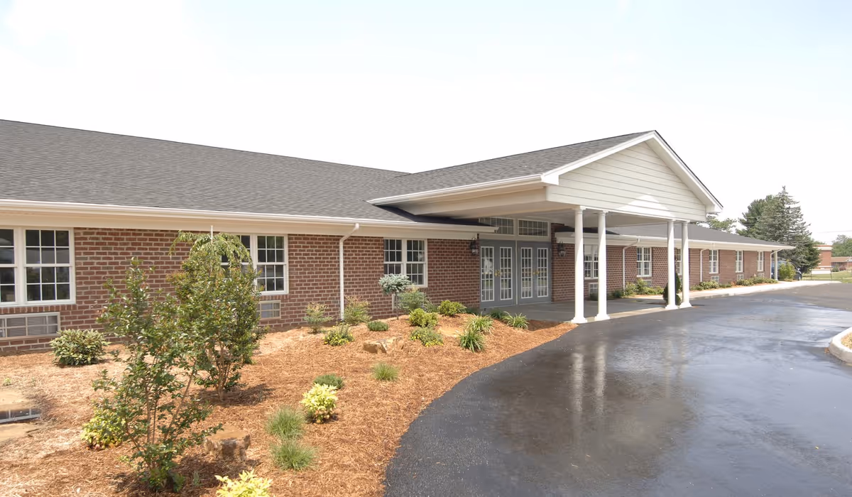 Exterior view of Shenandoah Place Assisted Living facility showing a single-story brick building with multiple windows, a covered entrance supported by white columns, landscaped garden beds with small shrubs and plants, and a paved driveway.