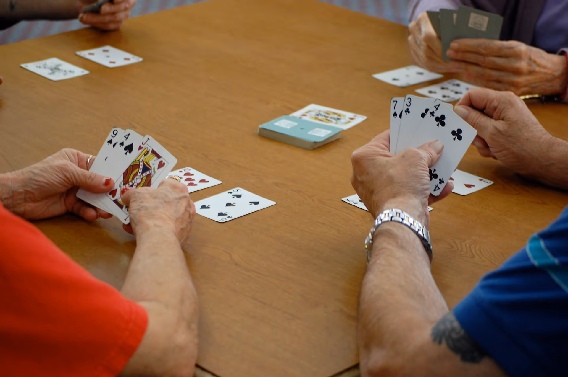 Four elderly people sitting around a wooden table playing a card game, each holding playing cards in their hands with some cards laid out on the table.