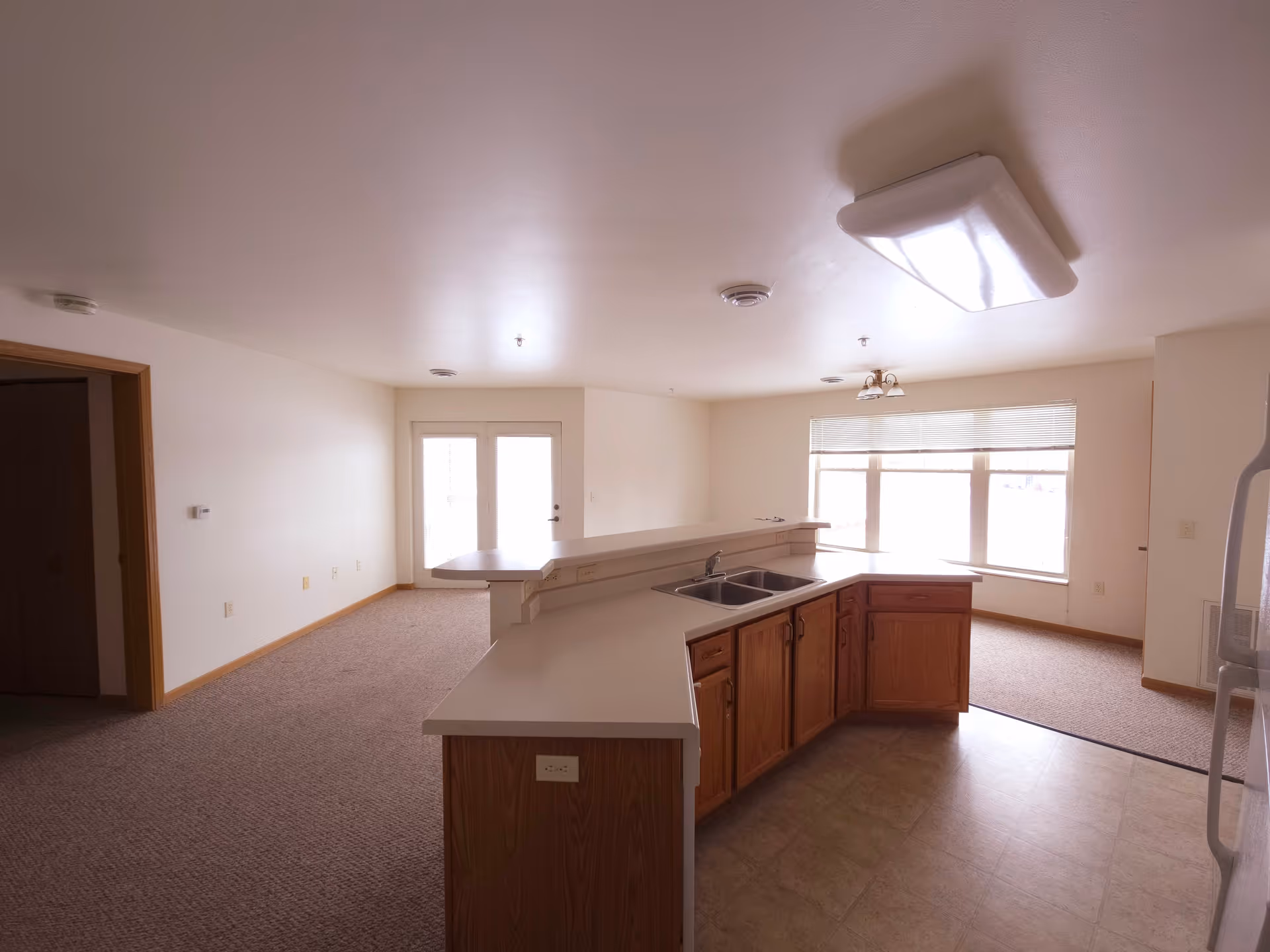 Interior view of a senior living facility unit showing a kitchen area with wooden cabinets, a double sink, and a countertop that extends into a small bar area. Adjacent to the kitchen is a carpeted living space with large windows letting in natural light and a door leading outside.