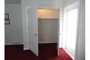 Interior view of a room with an open closet featuring double doors and a hanging rod inside. The room has dark red carpet flooring, light-colored walls, and a glass door on the right side allowing natural light to enter.