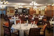 A spacious dining room with multiple round tables covered in white tablecloths, each set with folded napkins, glasses, and silverware. The room features wooden paneling, patterned upholstered chairs, chandeliers hanging from the ceiling, and a warm, inviting ambiance.