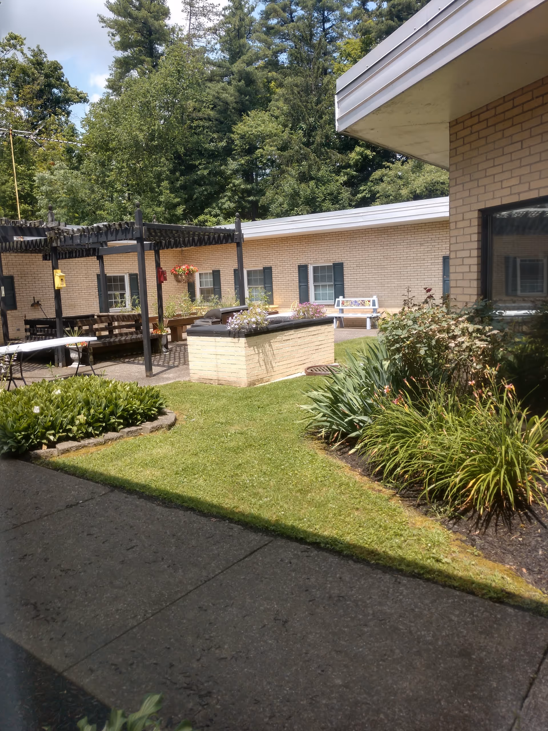 Outdoor courtyard area of a senior living facility with green grass, flower beds, and a brick building with windows and green shutters. There is a pergola structure with benches and tables underneath, and a white bench near the building. Trees are visible in the background under a partly cloudy sky.